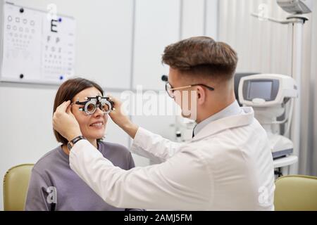 Ritratto di giovane oftalmologo mettere quadro di prova su paziente femminile sorridente durante il controllo visivo in clinica moderna, copia spazio Foto Stock