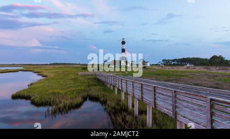 Tramonto al Faro di Bodie Island nelle Outer Banks, Carolina del Nord, con passerella in legno in primo piano sulla palude dell'oceano Foto Stock