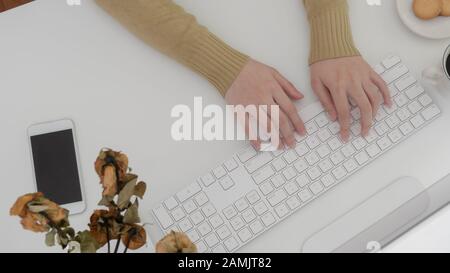 Vista dall'alto del freelancer femminile che digita sul computer su un moderno spazio di lavoro con biscotti, smartphone e rose secche su sfondo bianco scrivania Foto Stock