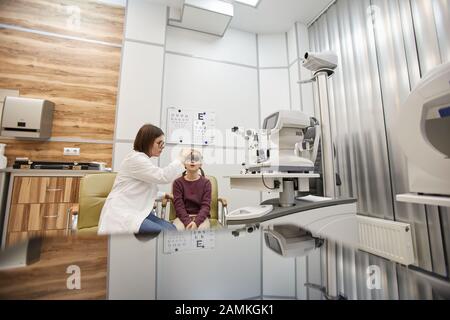 Ritratto grandangolare di cute ragazza teenage che indossa la cornice di prova durante il test di visione dei bambini in clinica oftalmologia moderna, copia spazio Foto Stock