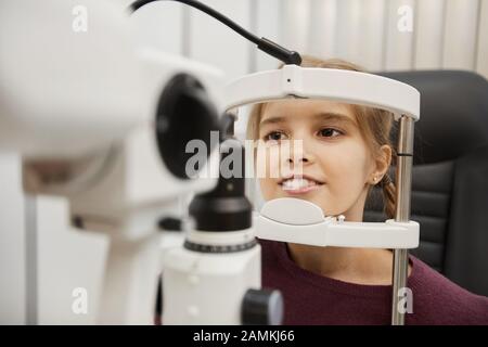 Ritratto di cute ragazza sorridente che guarda in rifrattometro durante il test di visione in moderna clinica oftalmologia, copia spazio Foto Stock