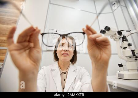 POV shot di giovane optometristo femminile che mette su occhiali su paziente irriconoscibile durante il test di visione in moderna clinica oftalmologia, copia spazio Foto Stock