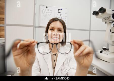 POV shot di sorridente optometristo femminile che mette sugli occhiali su un paziente irriconoscibile durante il test di visione in moderna clinica oftalmologia, copia spazio Foto Stock
