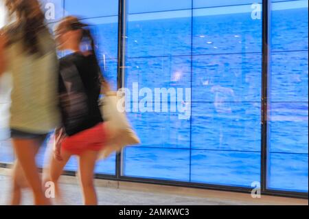 Due giovani donne che fanno una "passeggiata sulla spiaggia", se solo di fronte ad una proiezione video del mare nella vetrina del negozio *Hollister' nel Hofstatt di Monaco. [traduzione automatizzata] Foto Stock