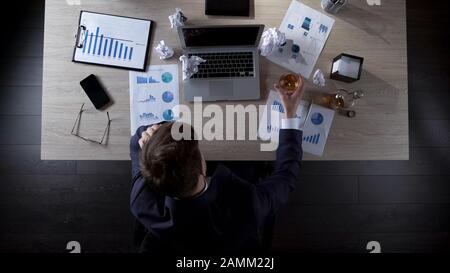 Uomo d'affari sconvolto seduto al tavolo e bere alcol, depressione, vista dall'alto Foto Stock