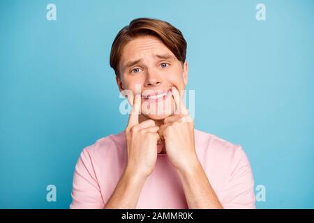 Ritratto di negativo infelice ragazzo rendendo sorriso sul suo volto con le dita l'usura alla moda vestito elegante isolate su blu colore di sfondo Foto Stock