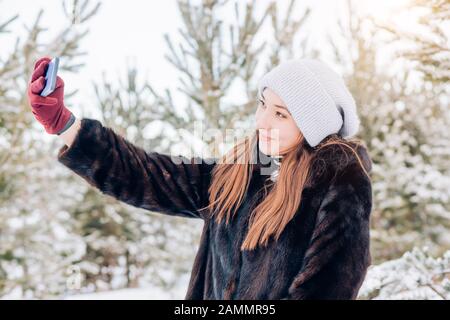 Closeup all'aperto ripresa di sorridente giovane turista femminile prende selfie. Donna caucasica che indossa un cappello a maglia, un cappotto in pelliccia calda e guanti sottili rossi Foto Stock