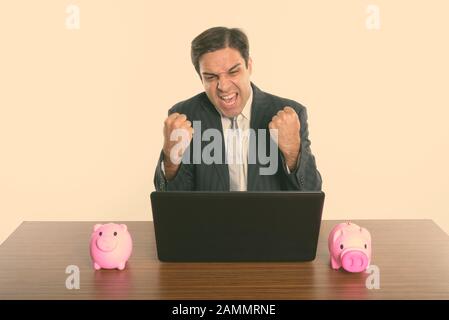 Un uomo d'affari persiano che guarda eccitato mentre usa un laptop con due banche piccanti su un tavolo di legno Foto Stock