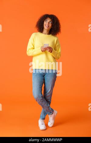 Full-length verticale scatto premuroso, bello e sognante africano americano donna con capelli ricci, guardando in su e sorridente, immagini cose, in piedi Foto Stock