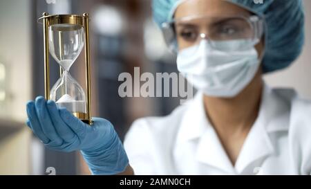 Scienziato femminile che guarda clessidra con sabbia che scorre giù, tempo limitato Foto Stock