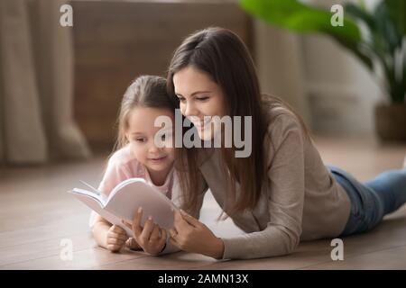 Libro di lettura sorridente della madre e della figlia piccola, giacente sul pavimento Foto Stock