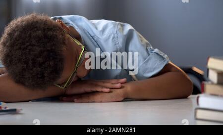 Ragazzo non motivato della scuola che dorme sulla scrivania della biblioteca vicino ai libri del palo, lezione noiosa Foto Stock