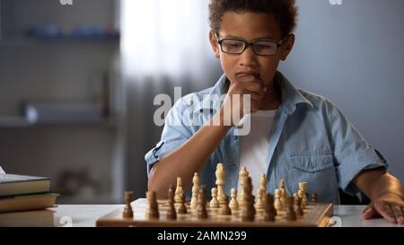 Piccolo ragazzo che pensa al movimento degli scacchi, hobby intelligente, sviluppo logico, tempo libero Foto Stock