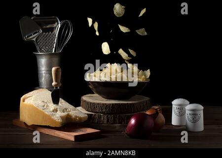 Patatine che cadono in una ciotola su un tavolo con parmigiano formaggio, cipolle, sale e pepe - umore scuro Foto Stock