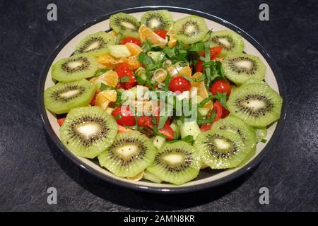 Un sano in casa ciotola di kiwi, pomodori ciliegini, cetriolo, foglie di lattuga, cipolline e mandarini sciroppati in insalata, Foto Stock