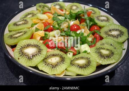 Un sano in casa ciotola di kiwi, pomodori ciliegini, cetriolo, foglie di lattuga, cipolline e mandarini sciroppati in insalata, Foto Stock