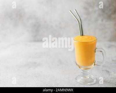 Giallo mango yogurt frullato o sul cemento grigio Sfondo con copia spazio a sinistra. Curcuma Lassie o lassi in vetro con cannucce di metallo Foto Stock