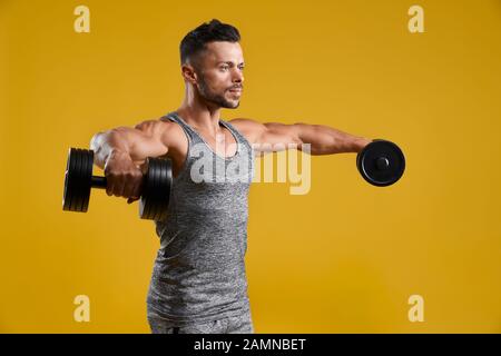 Vista laterale del gentiluomo muscolare di sollevamento pesi pesanti. Forte culturista in tank top fare esercizio con manubri. Isolato su giallo di sfondo per studio. Concetto di sport e di duro allenamento. Foto Stock