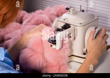 Master professionale per la produzione di capi di abbigliamento da pelliccia. Donna degno alla macchina da cucire cuce i vestiti da pelliccia rosa. Il processo di cucitura Foto Stock