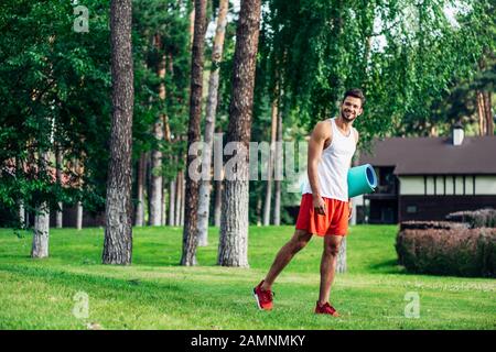 allegro uomo con bearded che tiene tappeto fitness mentre cammina nel parco Foto Stock