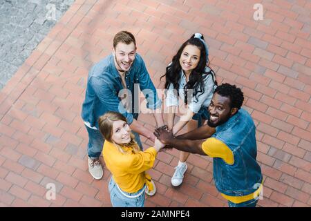 vista dall'alto di amici multiculturali allegri che mettono insieme le mani e guardano la macchina fotografica Foto Stock