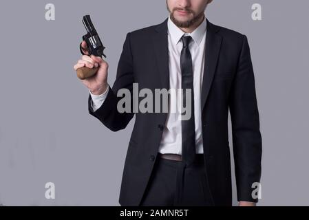 Ritratto di un uomo bearded in un vestito di affari che tiene un revolver. Su sfondo grigio. Uomo di tipo criminale, gangster, killer, uomo d'affari killer Foto Stock