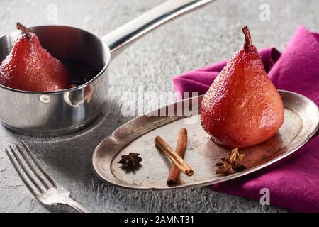 deliziosa pera al vino con cannella e anice su piastra d'argento e in stufato su superficie di cemento grigio con tovagliolo e forchetta rosa Foto Stock