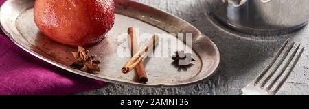 deliziosa pera al vino con cannella e anice su piastra d'argento e in stufato su superficie di cemento grigio con tovagliolo e forchetta rosa, colpo panoramico Foto Stock