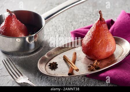 deliziosa pera al vino con cannella e anice su piastra d'argento e in stufato su superficie di cemento grigio con tovagliolo e forchetta rosa Foto Stock