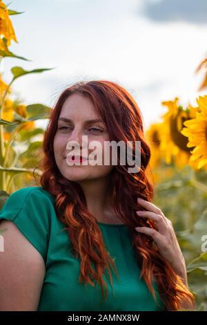 Bella ragazza giovane con capelli ricci rossi e un abito verde che posano in un fieldon girasoli una giornata di sole in estate Foto Stock