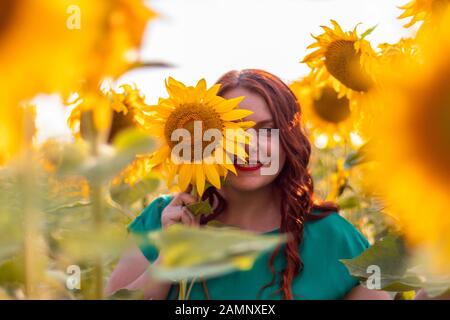 Ritratto di una bella ragazza asiatica con capelli ricci rossi e un abito verde che posa in un campo di girasoli in estate in una giornata di sole. Libertà Foto Stock