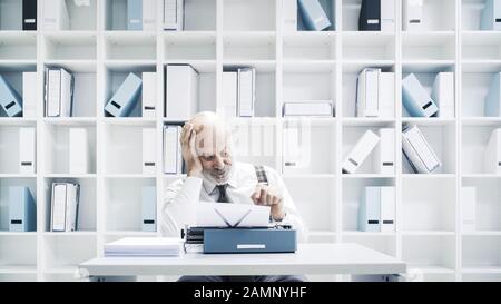 Uomo d'affari pigro senior o giornalista che lavora in ufficio, è seduto alla scrivania e utilizzando una macchina da scrivere Foto Stock