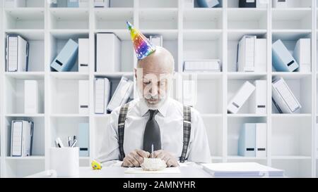 Uomo d'affari senior che ha un triste compleanno solitario in ufficio, sta indossando un cappello da festa e avendo una piccola torta da solo Foto Stock