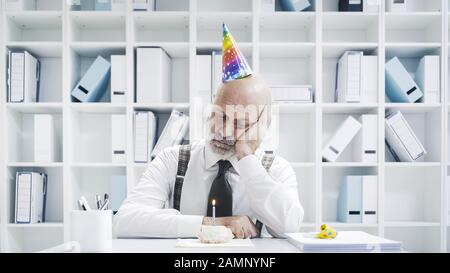 Uomo d'affari senior che ha un triste compleanno solitario in ufficio, sta indossando un cappello da festa e avendo una piccola torta da solo Foto Stock