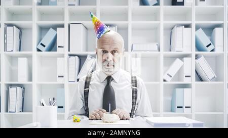 Uomo d'affari senior che ha un triste compleanno solitario in ufficio, sta indossando un cappello da festa e avendo una piccola torta da solo Foto Stock