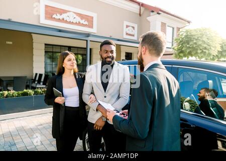 Giovane commerciante di automobile del caucasico che spiega il contratto di vendite per accoppiare nell'usura di affari, l'uomo africano e la donna caucasica, comprante un automobile, levandosi in piedi all'aperto Foto Stock