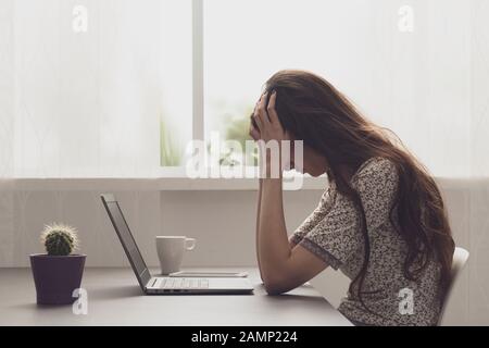 Stanco stressato donna seduta alla scrivania con la testa in mano e lavorando con il suo laptop, è stressata e esausta Foto Stock