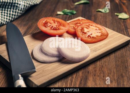 coltello con fette di pomodoro e cipolla su un tagliere e uno straccio su un tavolo di legno, cibo vegano e sano stile di vita Foto Stock