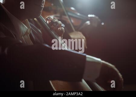 Orchestra sinfonica che si esibisce sul palco e suona un concerto di musica classica, violoncellista in primo piano, si avvicina Foto Stock