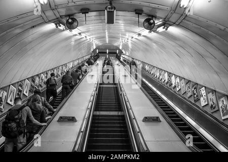 Vista monocromatica che guarda le scale mobili all'interno della stazione metropolitana di Londra. Vista posteriore in bianco e nero dei passeggeri del Regno Unito che viaggiano su una scala mobile. Foto Stock