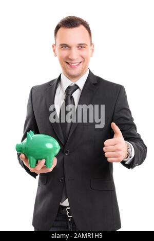 Bel giovane uomo d'affari in un vestito che tiene una banca piggy, sorridente e che dà il pollice in su, isolato su sfondo bianco. Risparmio di denaro. Foto Stock