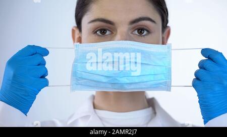 Giovane signora medico che mette sulla maschera chirurgica, la sicurezza durante l'esame, closeup Foto Stock