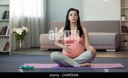 Donna incinta seduta su stuoia yoga fare esercizi di respirazione e massaggiare il ventre Foto Stock