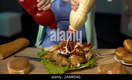 La ragazza obesa che versa il ketchup non sano e la maionese sul pollo arrosto grasso Foto Stock