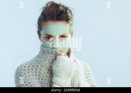 moderna casalinga di 40 anni in pullover roll collo e cardigan con maschera facciale verde coprente con vestiti isolati su inverno azzurro. Foto Stock