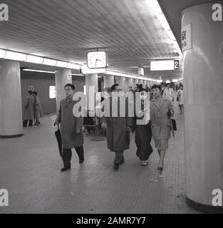 Anni sessanta, storico, Giapponese pendolari sulla Tokyo metro, con gli uomini in tute impermeabili e una donna giapponese che indossa un viso maschera dell'inquinamento. Foto Stock