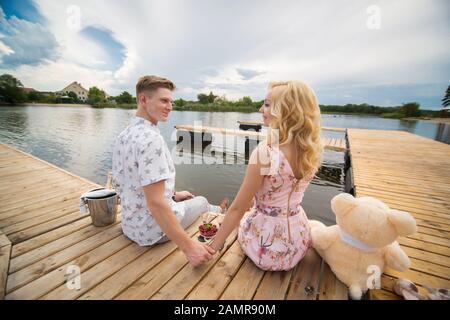 Data romantica sorpresa. Un ragazzo e una ragazza su un molo in legno. Il ragazzo versa dello Champagne nei bicchieri Foto Stock