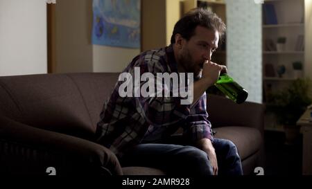 Uomo ubriaco seduto sul divano e bere birra in bottiglia, alcolismo e disperazione Foto Stock