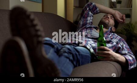 Ubriaco maschio dormire sul divano con bottiglia di birra in mano, cattive notizie, emozioni Foto Stock
