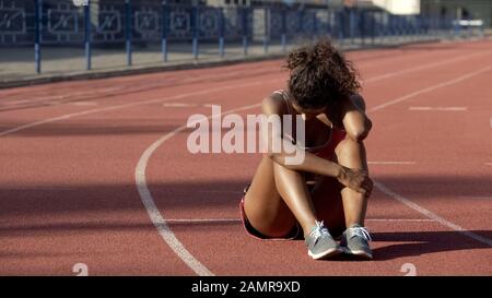 Sportswoman biracial esausto dopo allenamenti difficili seduti in mezzo alla pista Foto Stock